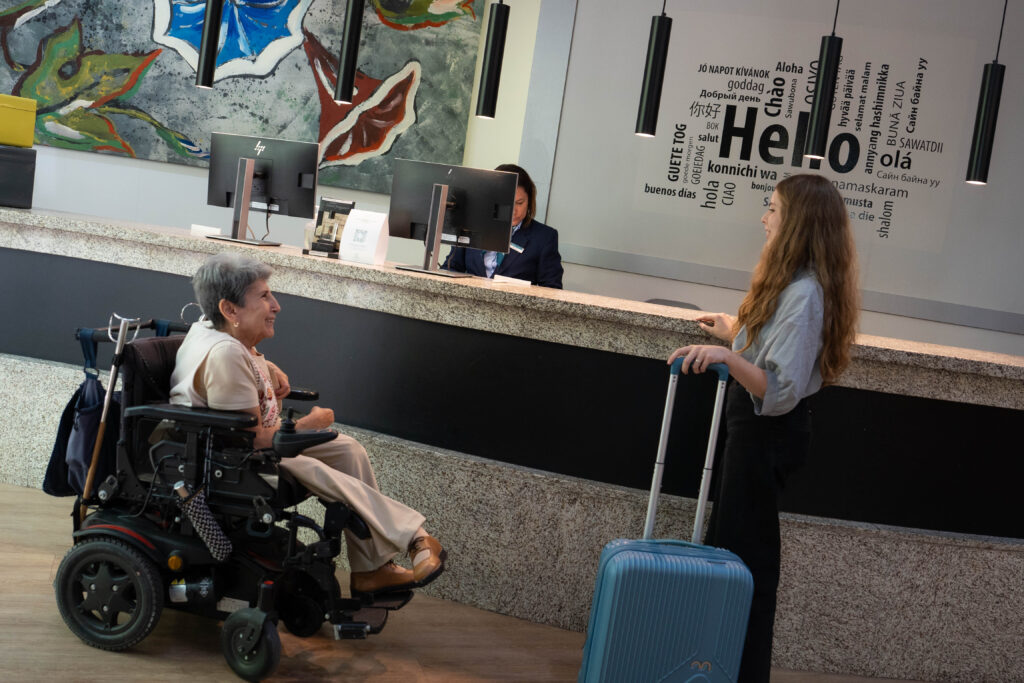 Una mujer en silla de ruedas hablando con otra mujer en un mostrador de recepción de hotel, ambas sonrientes. En la pared, se puede leer "Hello" en varios idiomas.