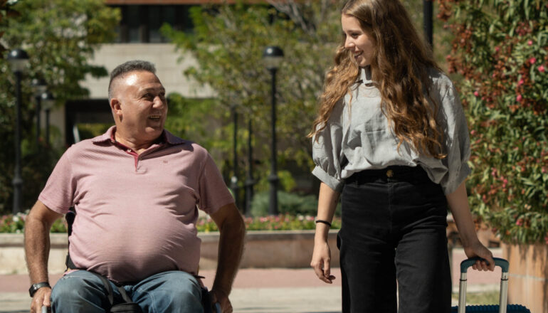 Un hombre en silla de ruedas y una mujer caminando juntos al aire libre en un entorno urbano. La mujer lleva una maleta con ruedas mientras conversan alegremente.