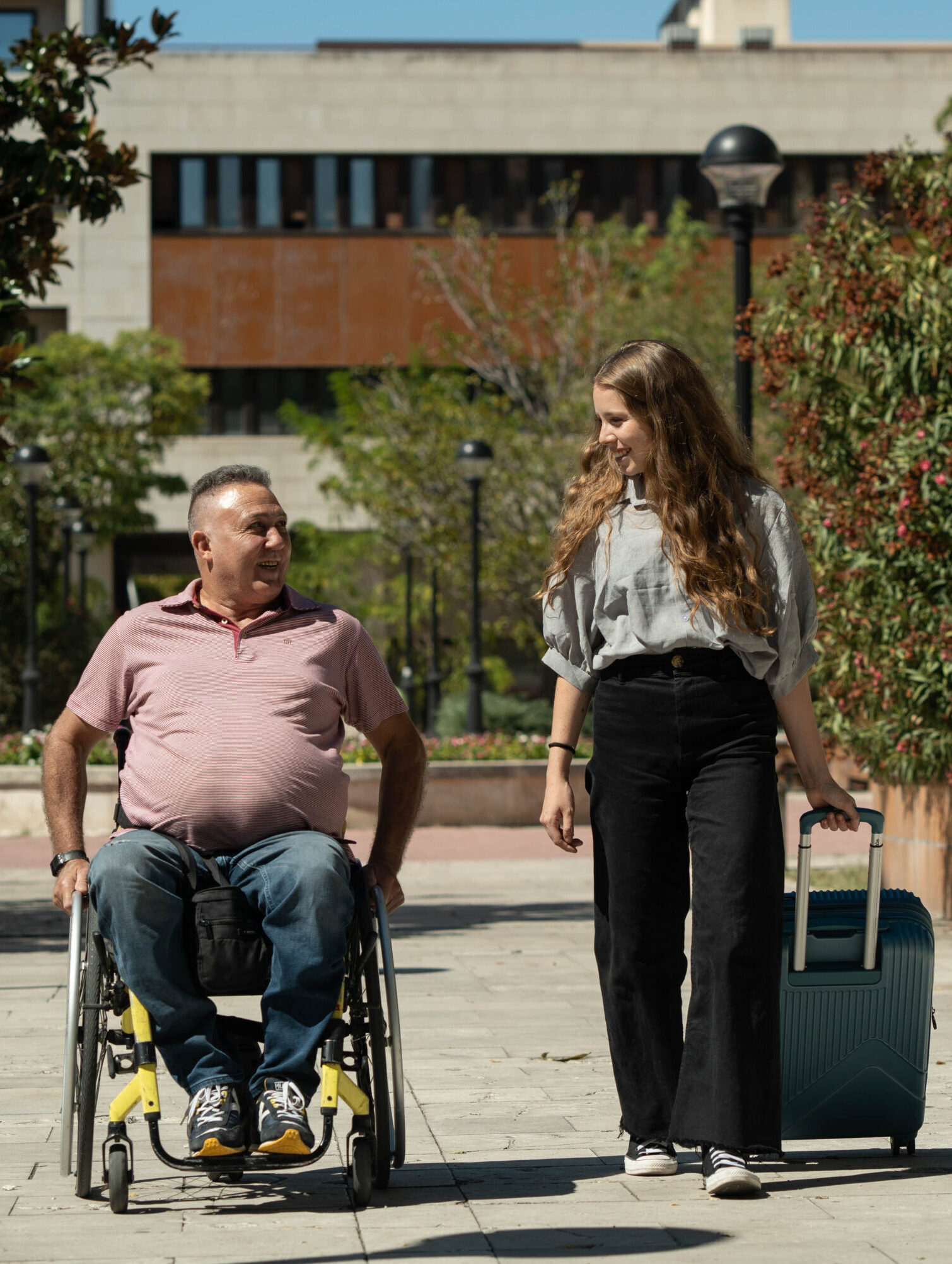 Un hombre en silla de ruedas y una mujer caminando juntos al aire libre en un entorno urbano. La mujer lleva una maleta con ruedas mientras conversan alegremente.